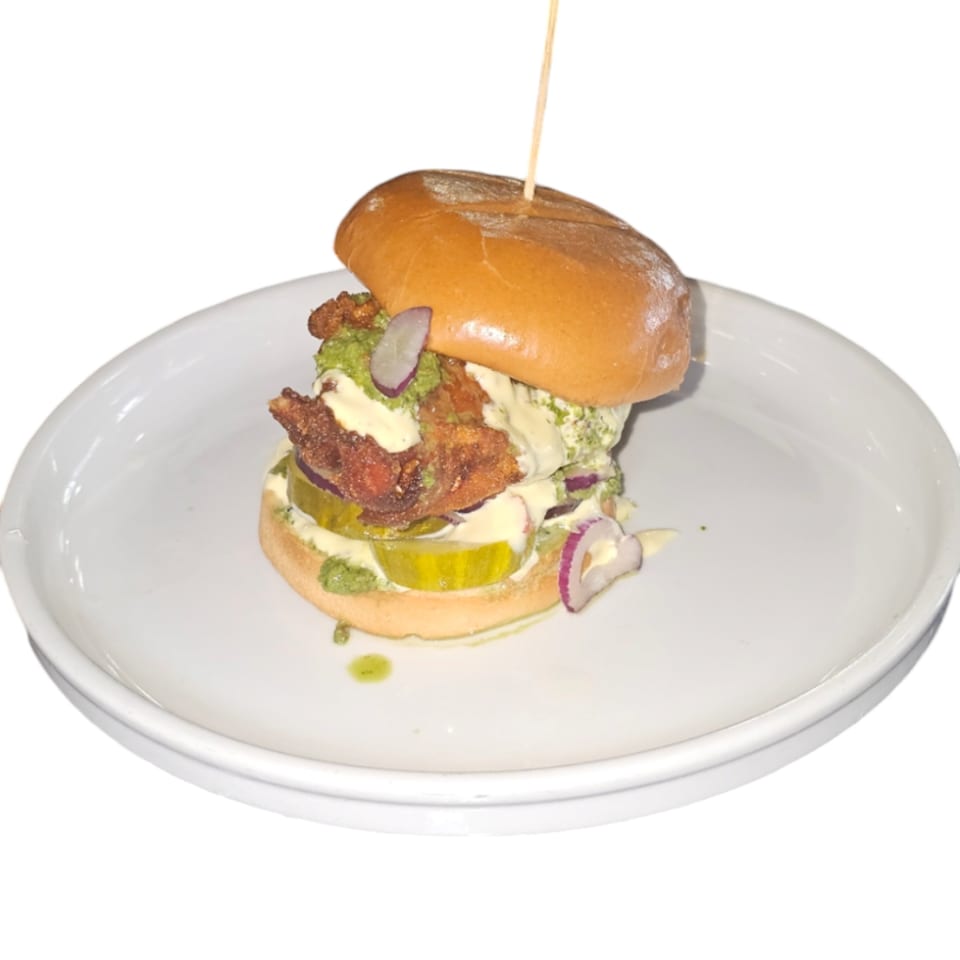 Halal fried chicken sandwich with pickles, red onion, and green sauce on a white plate.
