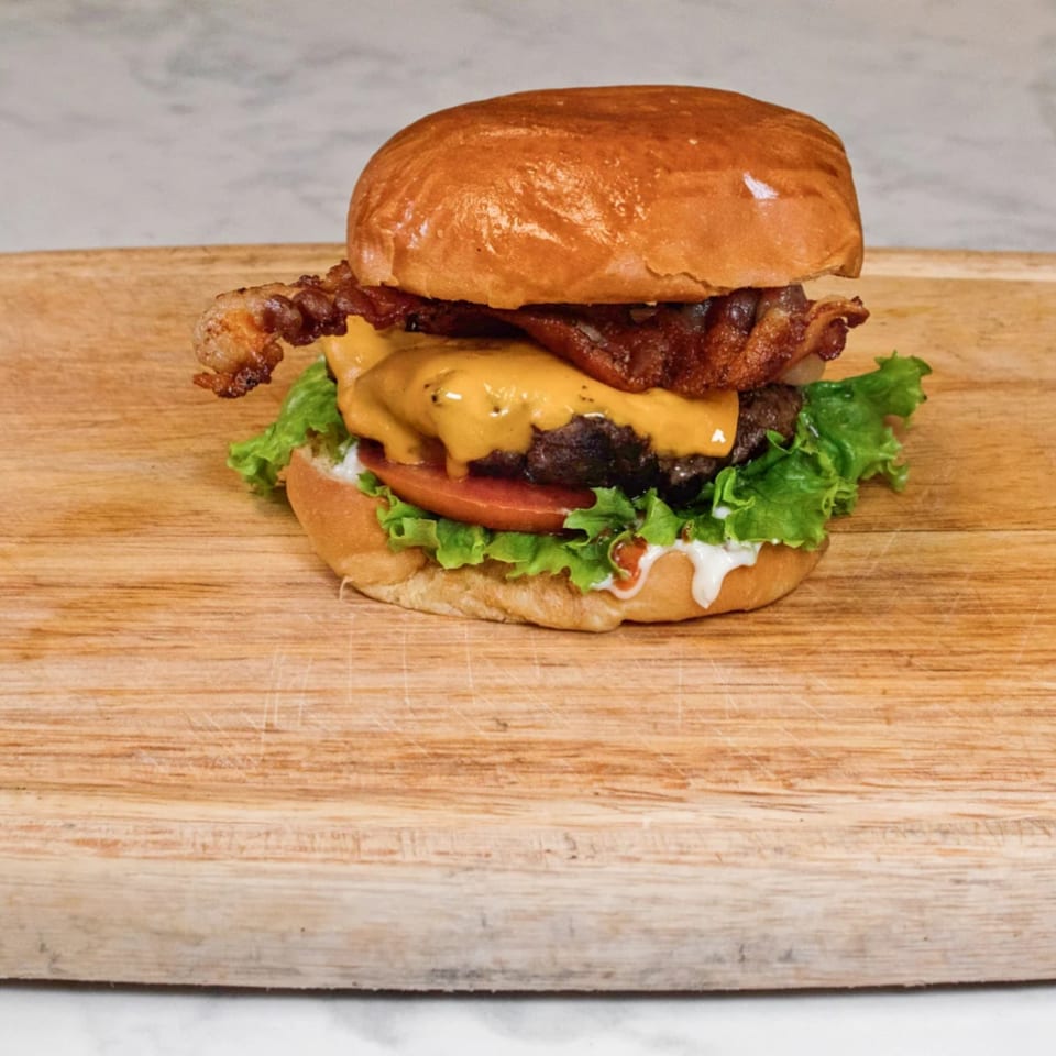 A halal burger with bacon, melted cheese, lettuce, tomato, and sauce on a wooden board.